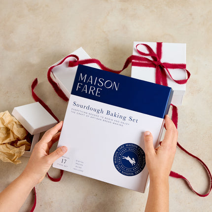 A person holds a Maison Fare Sourdough Baking Set box in front of several white gift boxes tied with red ribbon on a light beige surface.