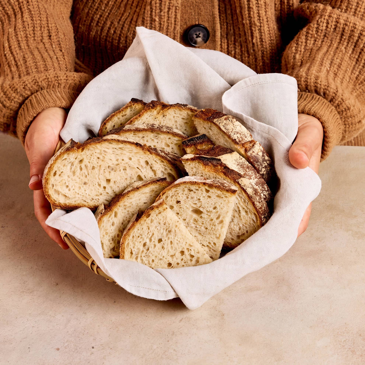 Slices of rustic artisan bread arranged in a woven basket lined with a white cloth, held by hands wearing a brown knit cardigan against a warm beige background.