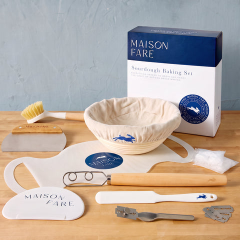 Maison Fare Sourdough Baking Set arranged on a wooden surface with its product box standing behind a beige flour-dusted bowl, white and wood tools, scraper, brush, and metal accessories against a pale blue wall.
