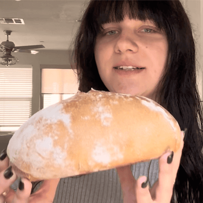 A person holding a large, rustic, flour-dusted bread loaf in front of their face in an indoor home setting.