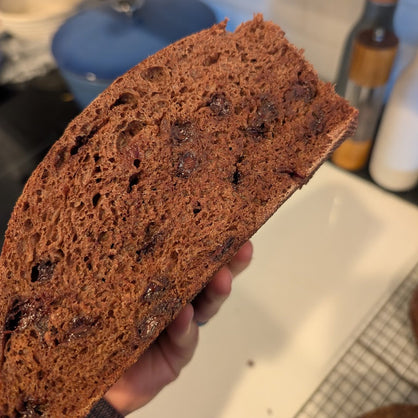 A close-up of a hand holding a thick slice of dark chocolate brown bread with visible dried fruit and chocolate chip pieces, set against a blurred kitchen background.