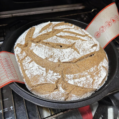 A round artisanal sourdough bread loaf with a flour-dusted crust and decorative slash marks, resting in a black cast iron skillet on a stovetop. Silicone sling handles stick out the sides. 