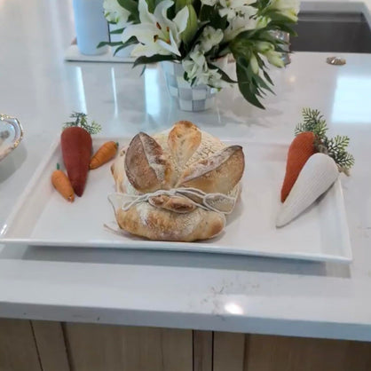 A rustic round sourdough bread loaf shaped like a bunny, tied with string on a white rectangular platter, styled with decorative carrots on a white kitchen countertop.
