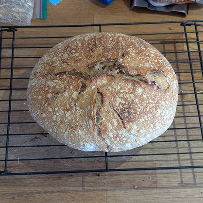 A round artisan sourdough bread loaf with a golden-brown crust sits on a black wire cooling rack over a wooden surface.
