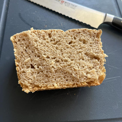 A thick slice of rustic whole wheat bread with a porous, airy crumb, resting on a dark cutting board next to a serrated knife.